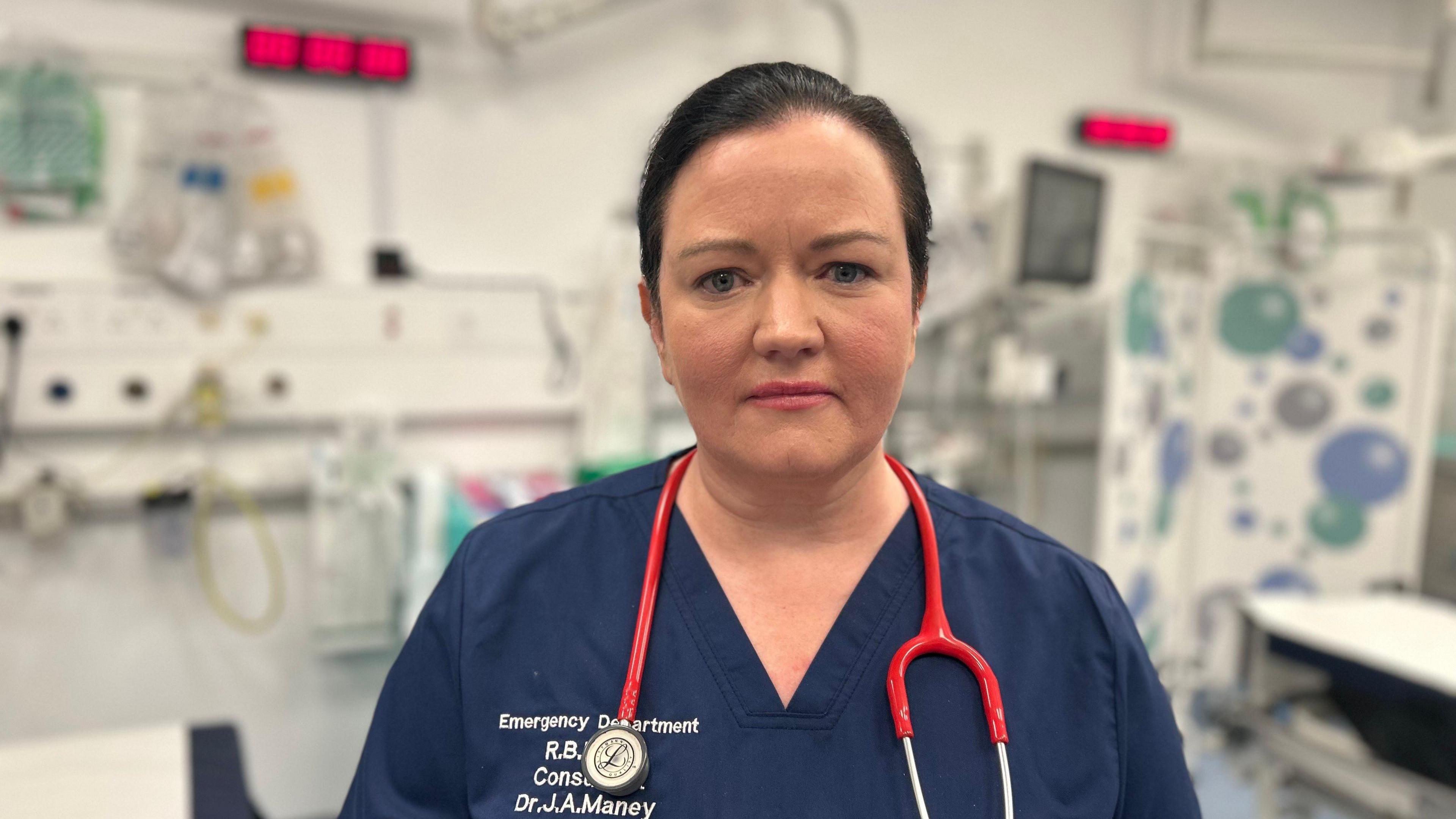 A shot of a woman wearing navy medical scrubs. She has short black hair and has a stethoscope around her neck. She is standing in a hospital room which is out of focus behind her.