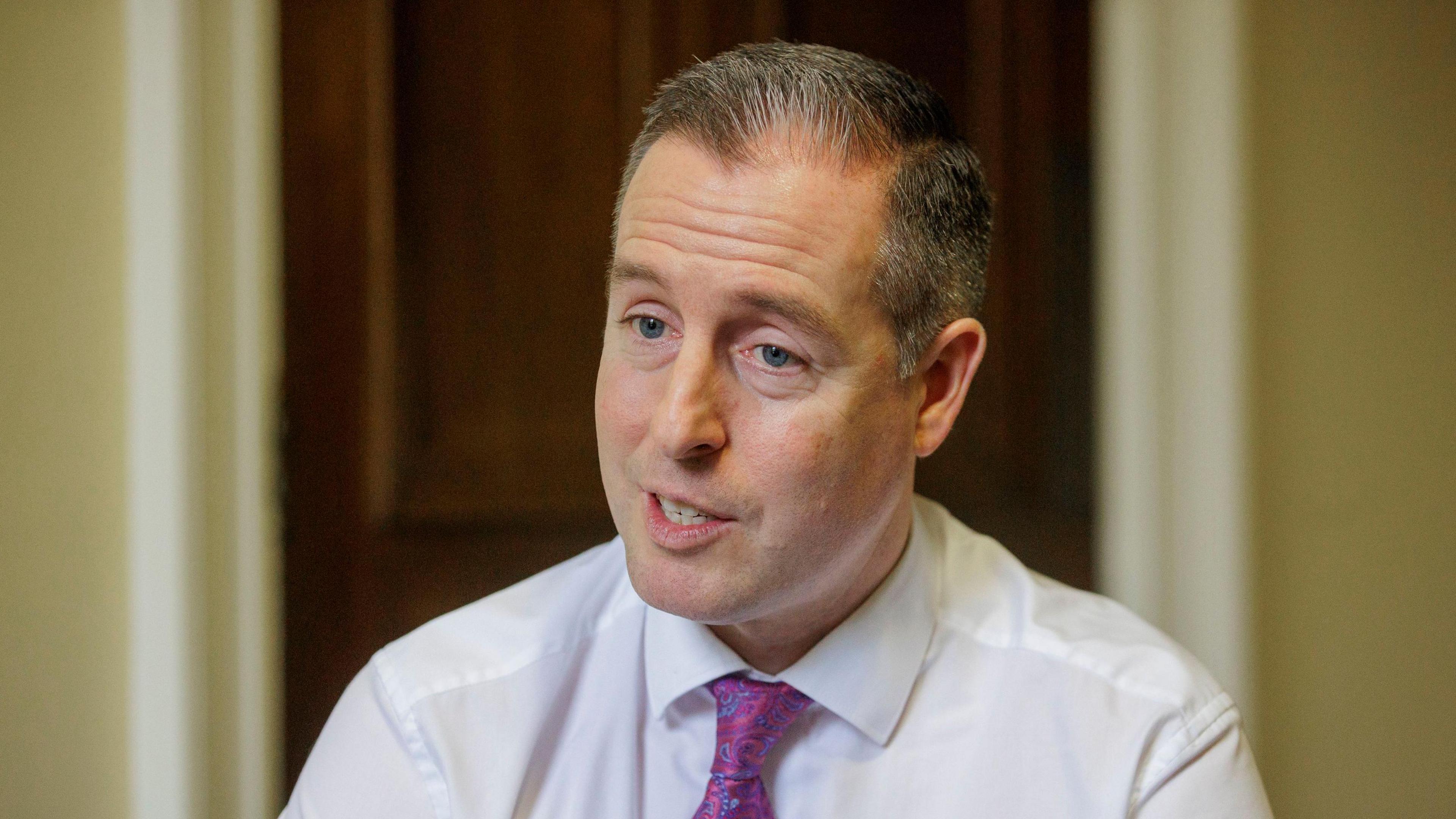 Paul Givan. He has shirt grey-ish hair and blue eyes, wearing a white shirt and purple paisley tie. A brown wooden door and yellow wall is blurred in the background.