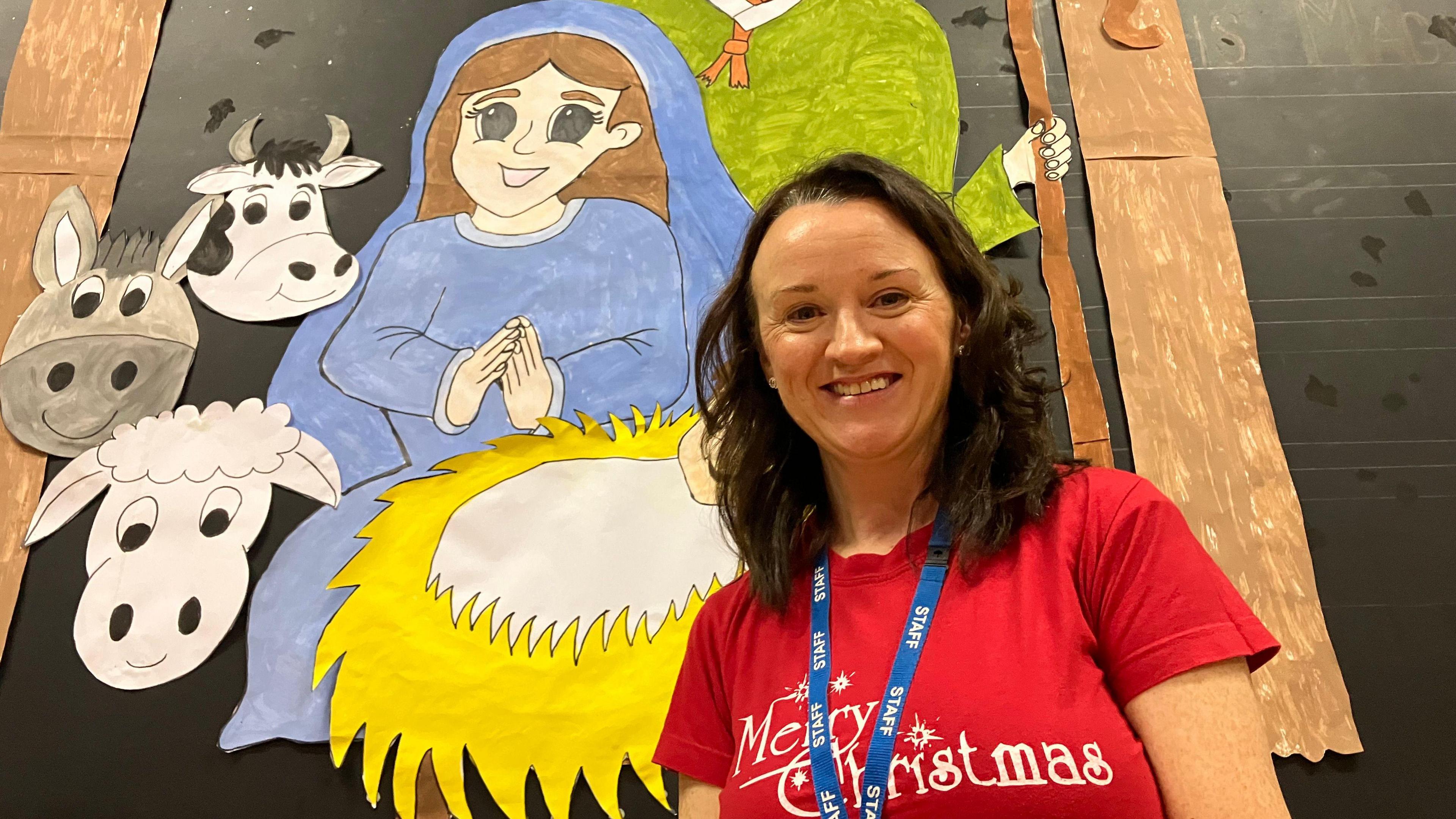 A dark haired woman with a merry christmas red t shirt smiles in front of a nativity scene