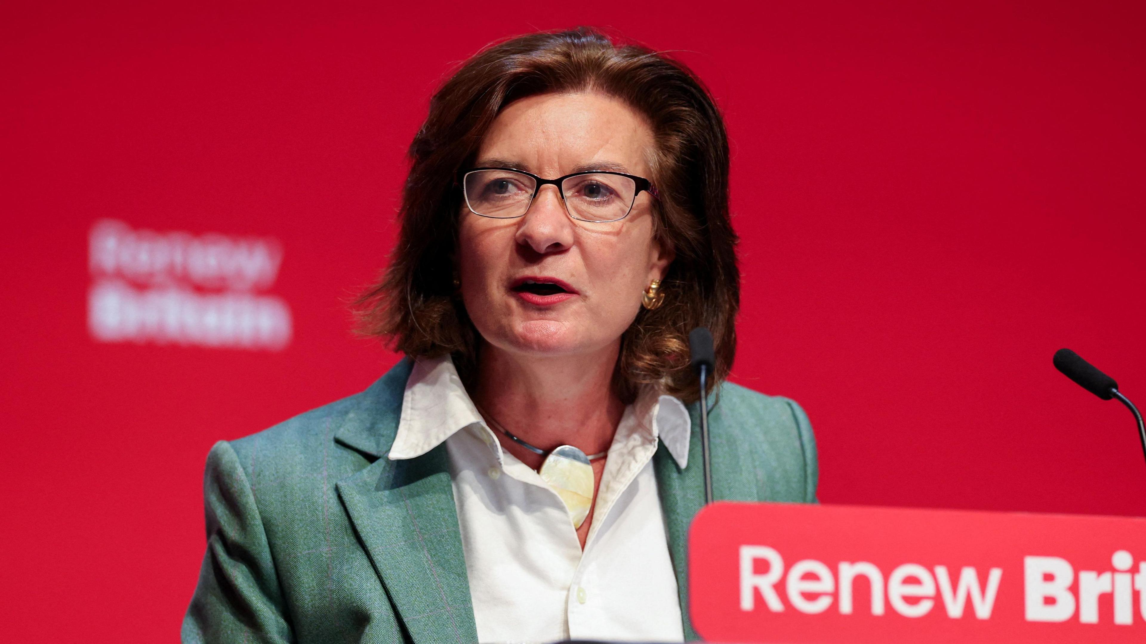 Eluned Morgan stood in front of a lecturn which has the words "Renew Britain" written across it but slightly out of view. She is wearing a green jacket and a white blouse, earrings and glasses.