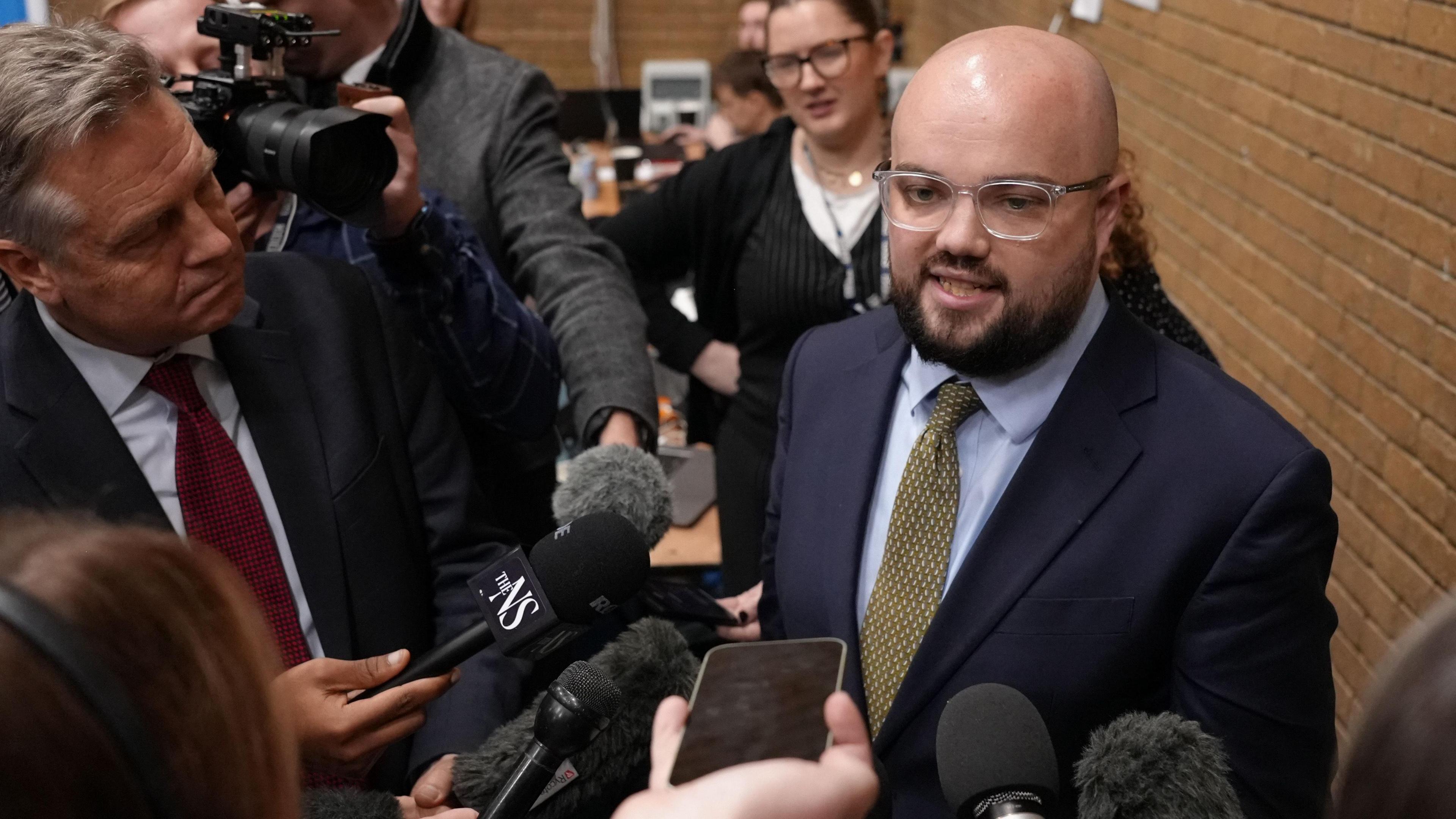 Llyr Powell speaking to reporters, with a brick wall behind him. Reporters are holding microphones and mobile phones in front of him.
