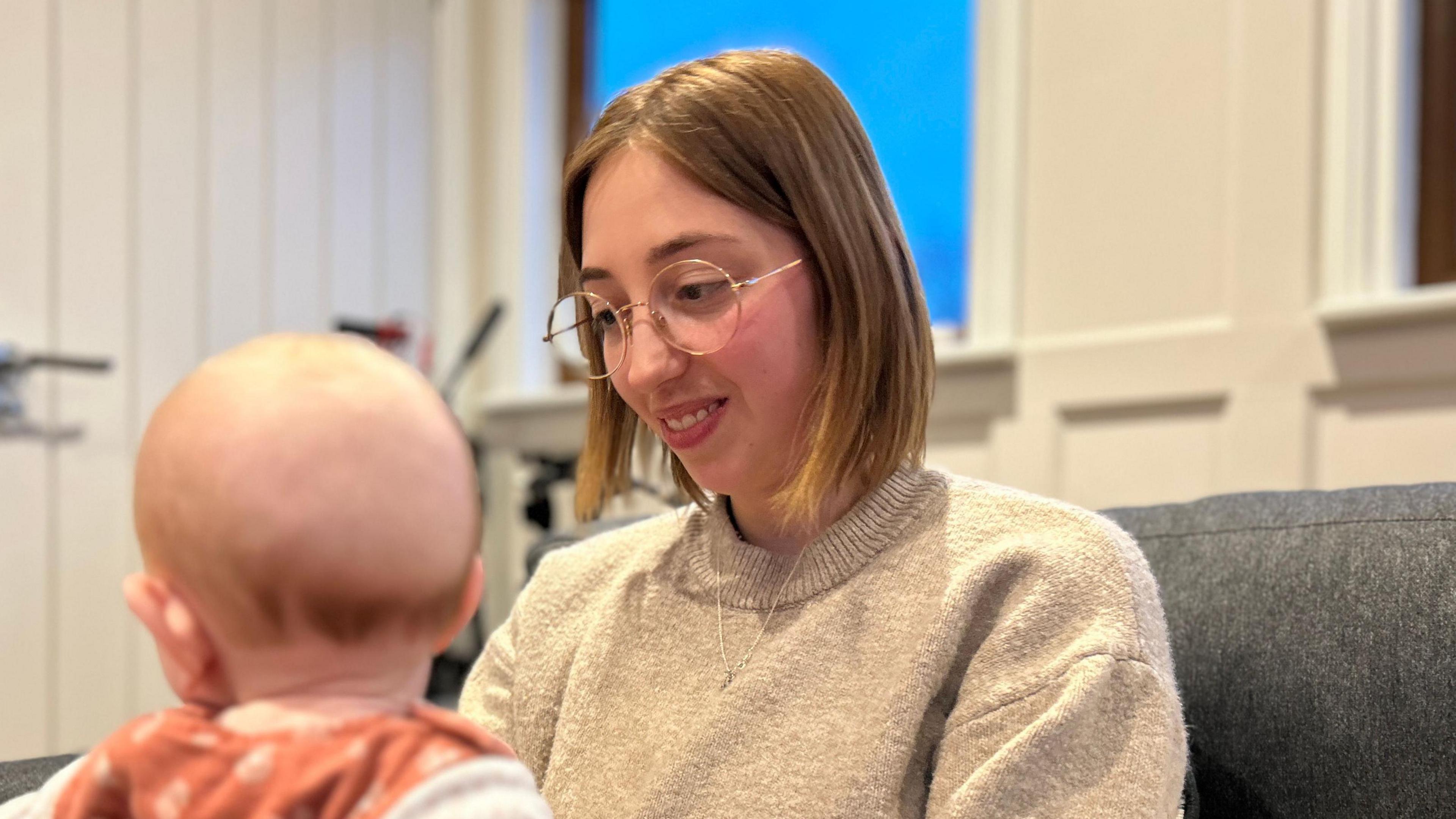 Woman with auburn bob hair smiling at a baby on her knee. She is wearing glasses and a beige wool jumper.