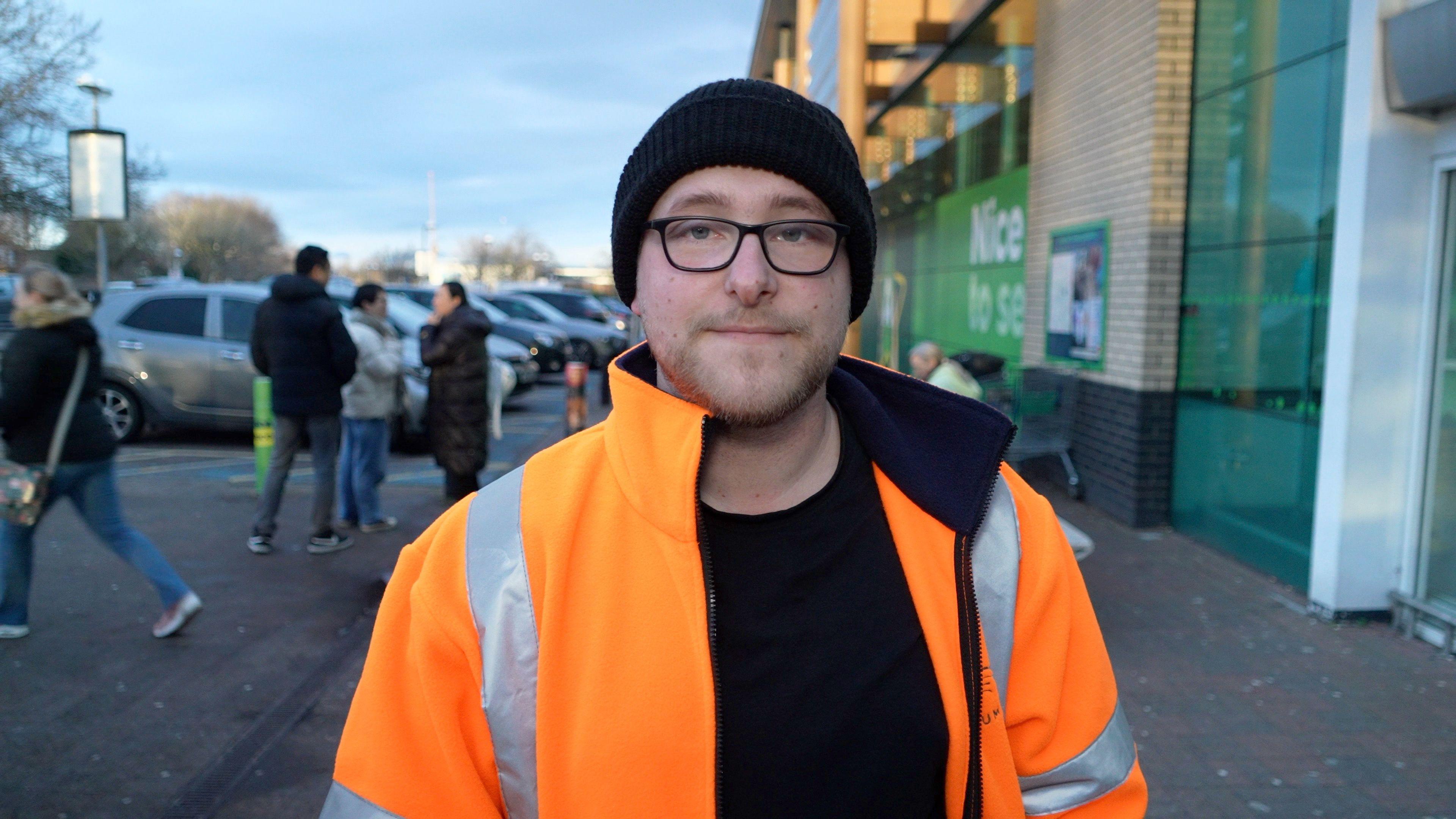 Jordon Elis looks expressionless at the camera. He is wearing a black jumper with orange high-visibility jacket on top. He is also wearing a black hat and black-rimmed glasses.