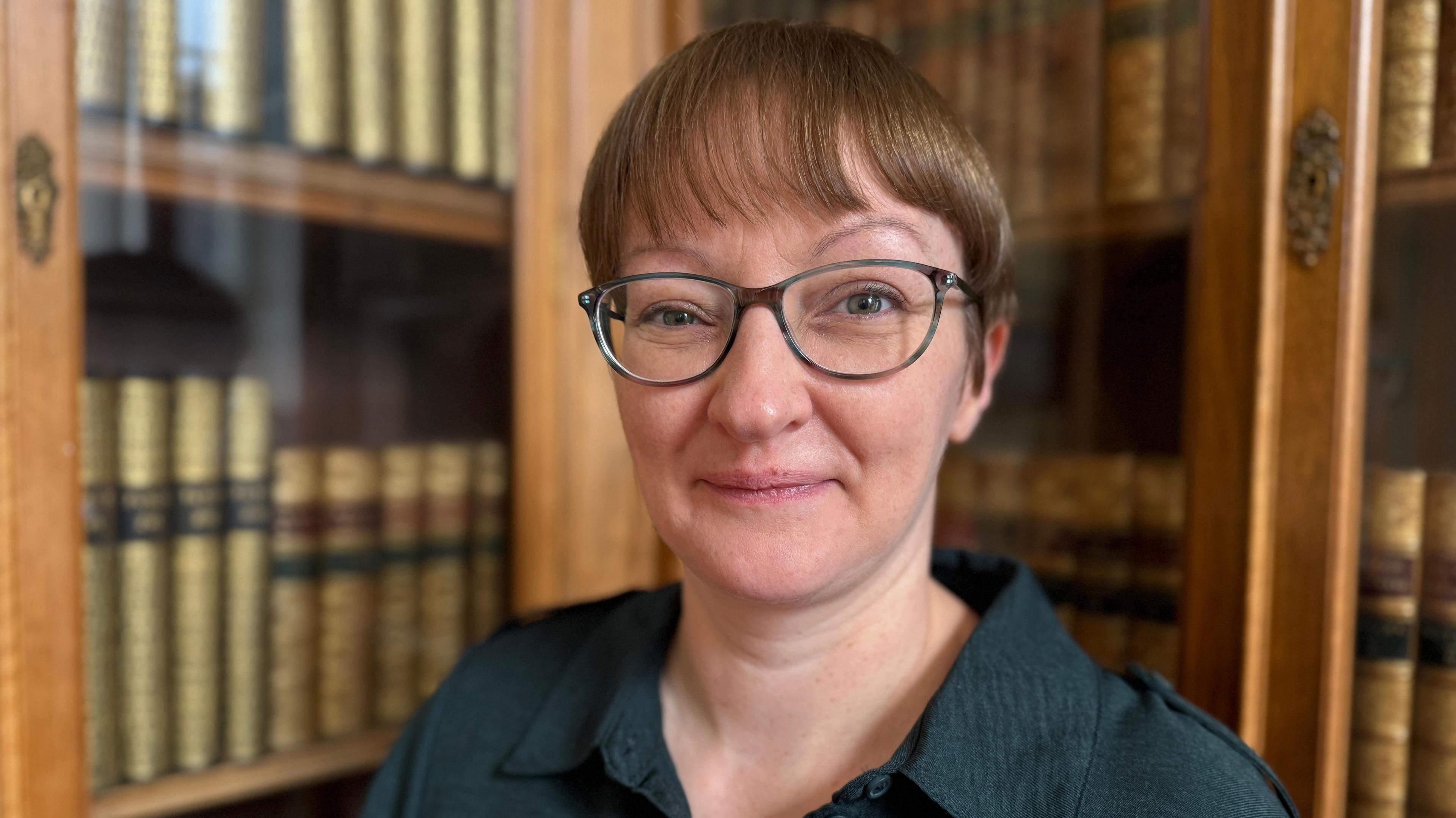 Shona Hunter standing in front of bookcases filled with books. She has short brown hair and glasses and is wearing a green shirt.