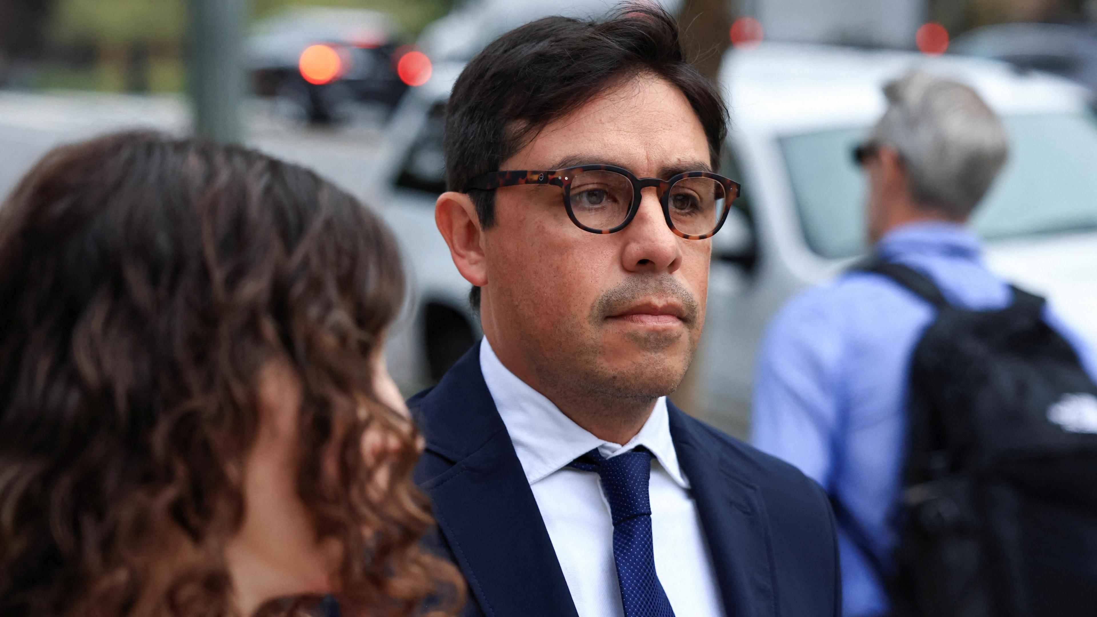 Dr Salvador Plasencia wears a suit and tie as he makes his way to federal court on 23. July 23, 2025. A person with long curly hair stands next to him.