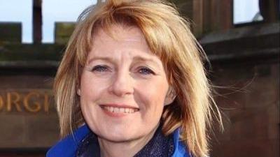 A woman with reddish fair hair is wearing a blue coat and darker blue scarf. She is standing in front of the red sandstone remains of Coventry Cathedral