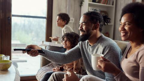 A family sitting down to watch TV, with a man holding a TV remote and woman holding a mug, the children are in the background