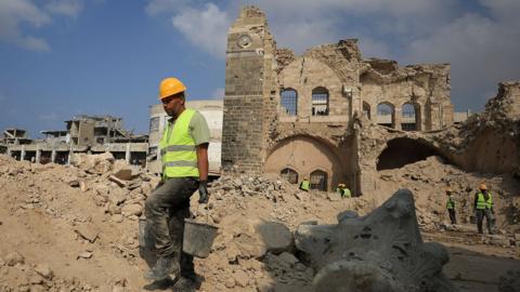 A worker in a high vis vest and yellow hard hat carries grey buckets while walking away from the site of Pasha's Palace in Gaza on 11 November.