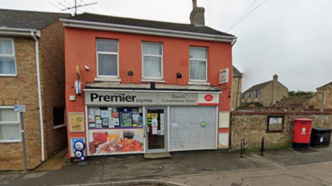 A red-painted two-storey detached building. The ground floor is a Post Office and Premier Express convenience store. To the right is a red post box on the pavement in front of a bricked wall, which has a noticeboard on it.