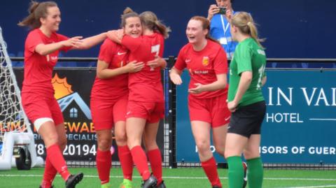 Jersey player dressed in red hug whilst a Guernsey player in green looks on. They are near the goal.