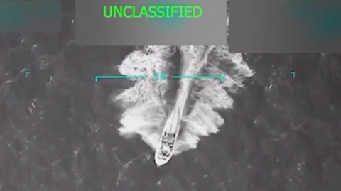 A grainy aerial video still shows a motor boat speeding across choppy waters