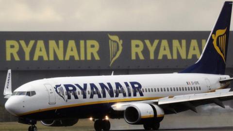 A Ryanair aeroplane taxiing down a runway. It is white with yellow and blue livery. Behind it is a large grey Ryanair hangar.