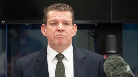 Rhun ap Iorwerth speaking into a microphone. He wears a dark suit, green patterned tie and white shirt.