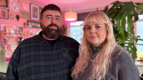 A man with dark hair and a blue and green tartan shirt stood next to a woman with blonde hair wearing a grey jumper.