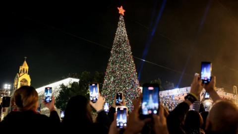 A giant, lit up Christmas tree with people holding phones standing around it