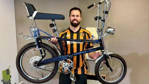 A man with slicked-back dark-brown hair and a beard smiles as he holds up a blue Raleigh Chopper bicycle against a plain sand-coloured wall. He is wearing a black and amber Hull City shirt. The bike is dark-blue with black wheels, a black seat, silver and black handlebars and silver trim.