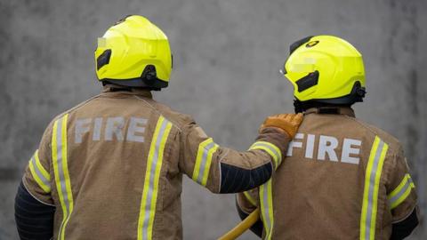 Two firefighters, wearing mainly brown clothing and yellow hard hats, have their backs to the camera. The one on the left wearing a brown glove has their right hand on the left shoulder of the colleague on the right.