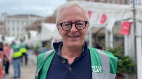A wide shot of Mike Powell. He has short grey hair and is wearing a dark green and grey Shelter Trust high visibility jacket.