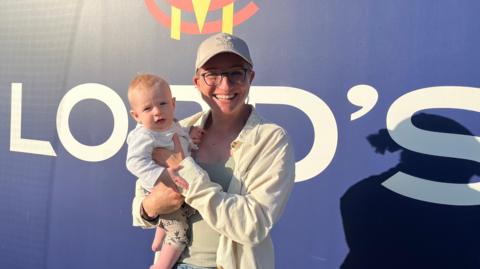 A woman wearing a cap and glasses stands in front of a sign at Lord's Cricket Ground in London, holding her infant son