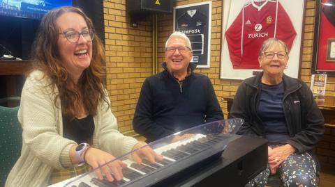 A woman is playing a keyboard, with a man and woman sat close beside her. The pianist has long brown hair, glasses, a cream woollen cardigan, a black top and a purple watch. The man is sat down wearing a black quarter zip fleece, glasses and has short white hair. The other woman also has glasses, a black jacket, navy top and navy leggings decorated with colourful dots. On the brick walls behind them are framed rugby shirts, a large black speaker and a large television. There are also two wooden tables behind them.