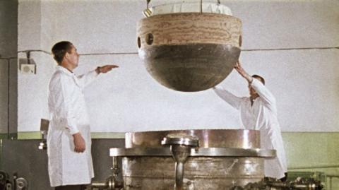 The image shows two individuals in white lab coats lowering the descent capsule of the Soviet space probe Venera 5 or 6 into a large, cylindrical metal structure to test the heat resistance. The capsule is spherical with a dark bottom half and a lighter top half, featuring several circular holes.