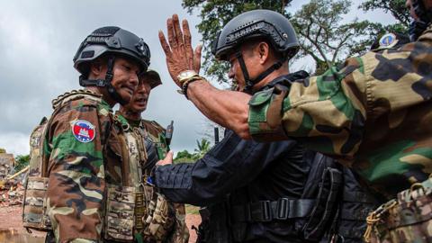 Two troops in Cambodian military uniform and helmets face off with a troop in black uniform and helmet, while in the foreground another soldier in military uniform holds out his hand with the palm acing outwards