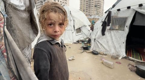 A Palestinian child, wet from rain, looks into the camera, surrounded by soaked tents and wet and muddy ground.