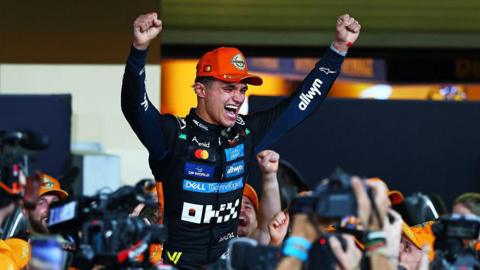 A racing driver dressed in a black racing suit with sponsors' badges all over the front. He has his hands aloft and is being supported on the shoulders of those in crowd.