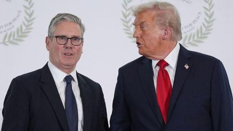 Sir Keir Starmer stands beside US President Donald Trump at an event, both wearing dark suits and ties, with Trump turned toward Starmer as if speaking while Starmer looks toward the camera.