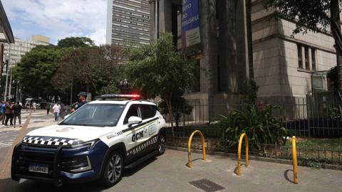 A police car with flashing lights stands in front of the main entrance to the Biblioteca Mario de Andrade in São Paulo. A uniformed police officer is standing guard next to the entrance. A banner hanging from the column at the left-hand side of the entrance advertises the exhibition "From book to museum".