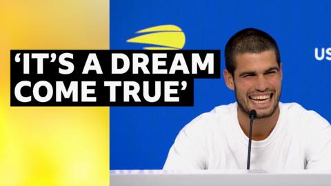 Tennis player Carlos Alcaraz smiling at a press conference after winning the US Open. He is wearing a white shirt while sitting at the presser desk with a blue background behind him.