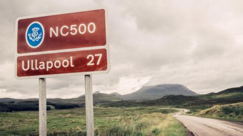A brown NC500 sign post which says it is 27 miles to Ullapool next to a country road with hills in the background.