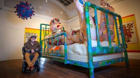 Artist Jason Wilsher-Mills sits alongside one of his exhibits - a giant inflatable character lying in a hospital bed watching TV inside the Grundy Art Gallery.