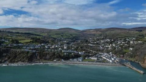An arial view of Laxey bay, you can see the coastline, the sea is turquoise, you can see houses scattered on a hilly green terrain.