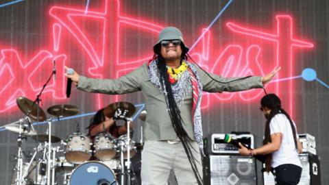 Maxi Priest, who has long dreadlocks and is wearing a patterned scarf and bucket hat, has his arms outstretched on stage. he has a microphone in one hand and there is a drummer and a guitarist in the background.