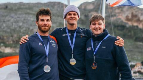 Three men stand in a row with their arms around each other. One is wearing a bronze medal, one silver and one gold. The man on the left has brown hair and a beard, the man in the centre is wearing a light purple beanie hat, and the man on the right has brown hair. All three are wearing navy-coloured long sleeve tops.