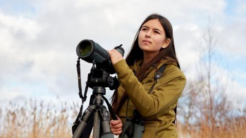 Dr Mya-Rose Craig holds on to a black telescope. She is looking off into the distance on an autumnal day. She has long brown hair and is wearing a brown jacket.