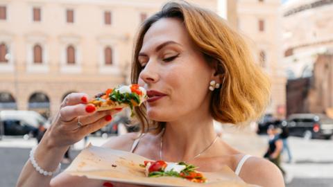 A woman eats a slice of pizza on the street in Rome. She has short hair.