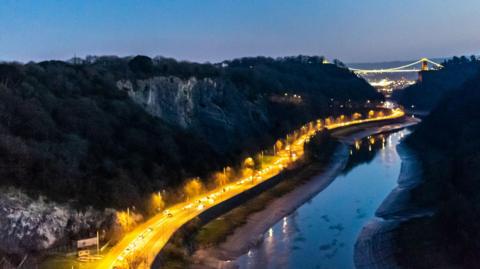 A night-time view of the Bristol Portway with traffic heading south. The road is brightly lit by street lights, making it stand out against the dark backdrop of the hills and river beside it. The suspension bridge can be seen in the background. Avon Gorge is to the left.