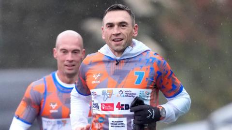 Kevin Sinfield running with a member of his support crew behind him. Sinfield is wearing an orange and blue top with the number 7 emlazoned on.
