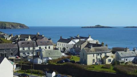 A view of houses against a backdrop of the sea. A set of houses, some detached. A cliff head can be seen in the background.