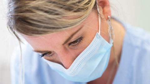 Close up of a blonde female nurse wearing a mask, dressed in light blue scrubs