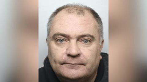 A mugshot of Green, who has short, thinning hair on the top of his head, with more hair visible on the sides. The hair appears to be a mix of grey and dark tones. He is wearing a dark-coloured hoodie, which is visible around the neck and shoulders. The background is plain and light-coloured.