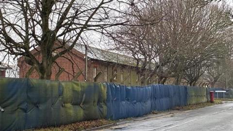 A tall fence with blue tarp running along it. There is a brick building and trees the other side of the fence.