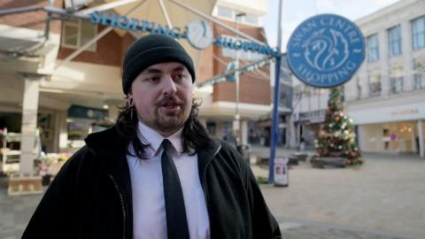 Man with a black hat, black jacket, white shirt, black tie outside a shopping centre