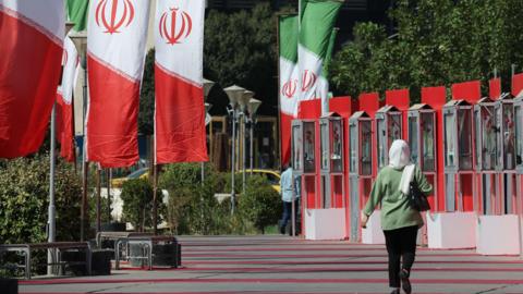 A woman in a green top, black pants and a white head scarf walks outside, away from the camera in the sun surrounded by Iranian flags. There are trees in the background.