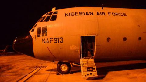 The nose of a Nigerian air force plan on the runway. The photograph is taken at night and the plane is bathed in an orange light.