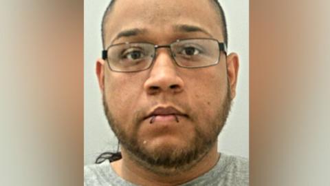 Robert Court's mugshot. He has dark hair and a short beard, black framed wire glasses and what looks like two big stitches through his bottom lip