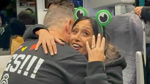 Steve Higgs, hugging Payal Pandya, you can only see his back. Payal has on green ears, on a hairband and is showing up her hand with a ring on it, looking excited. She has long dark hair.