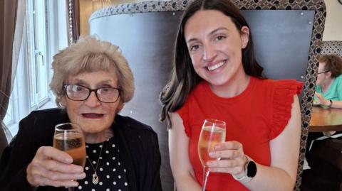 Two people sit at a restaurant table holding glasses of sparkling wine. One wears a red ruffled top, and has long dark brown hair Hannah. the other, Joan, is elderly and has shorter blowdried hair and glasses. Joan wears a black cardigan over a polka-dot top. Behind them is a tall booth with patterned fabric and round mirrors on the wall.